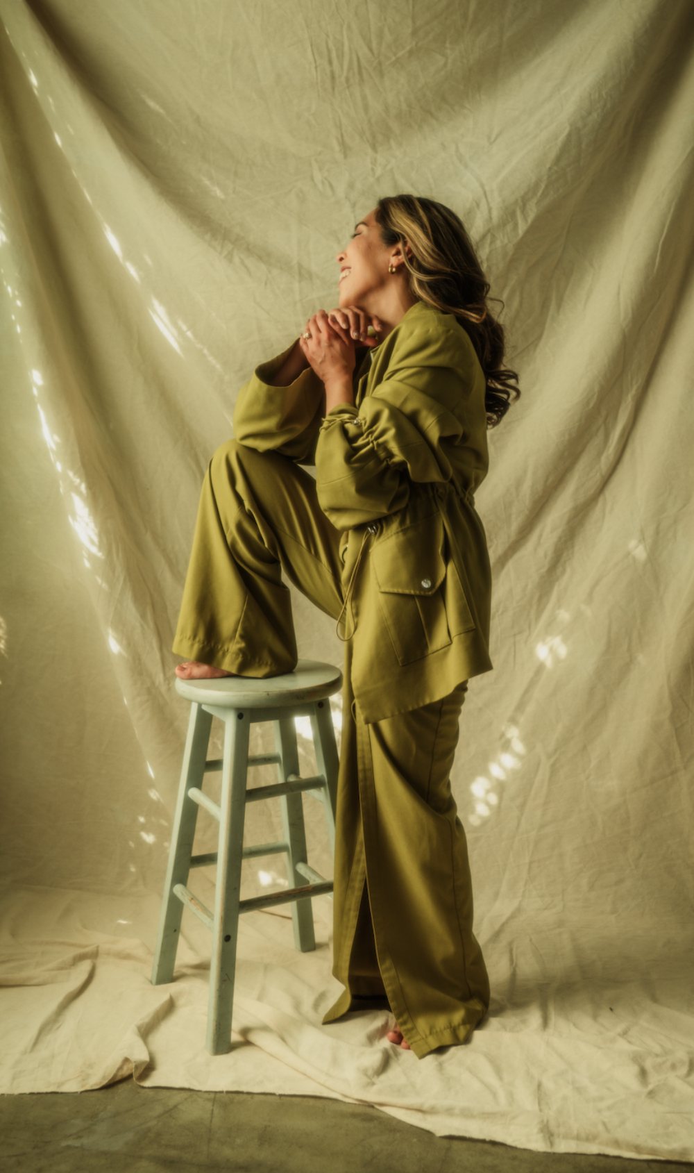 Charlene Platon smiling with one foot on a pale blue stool, in an olive suit against dappled linen