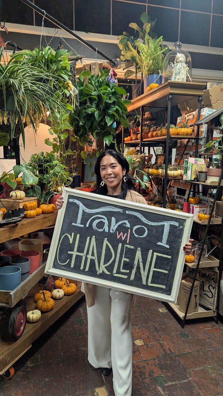 Charlene holding a chalkboard sign reading Tarot with Charlene in a pumpkin-filled plant shop