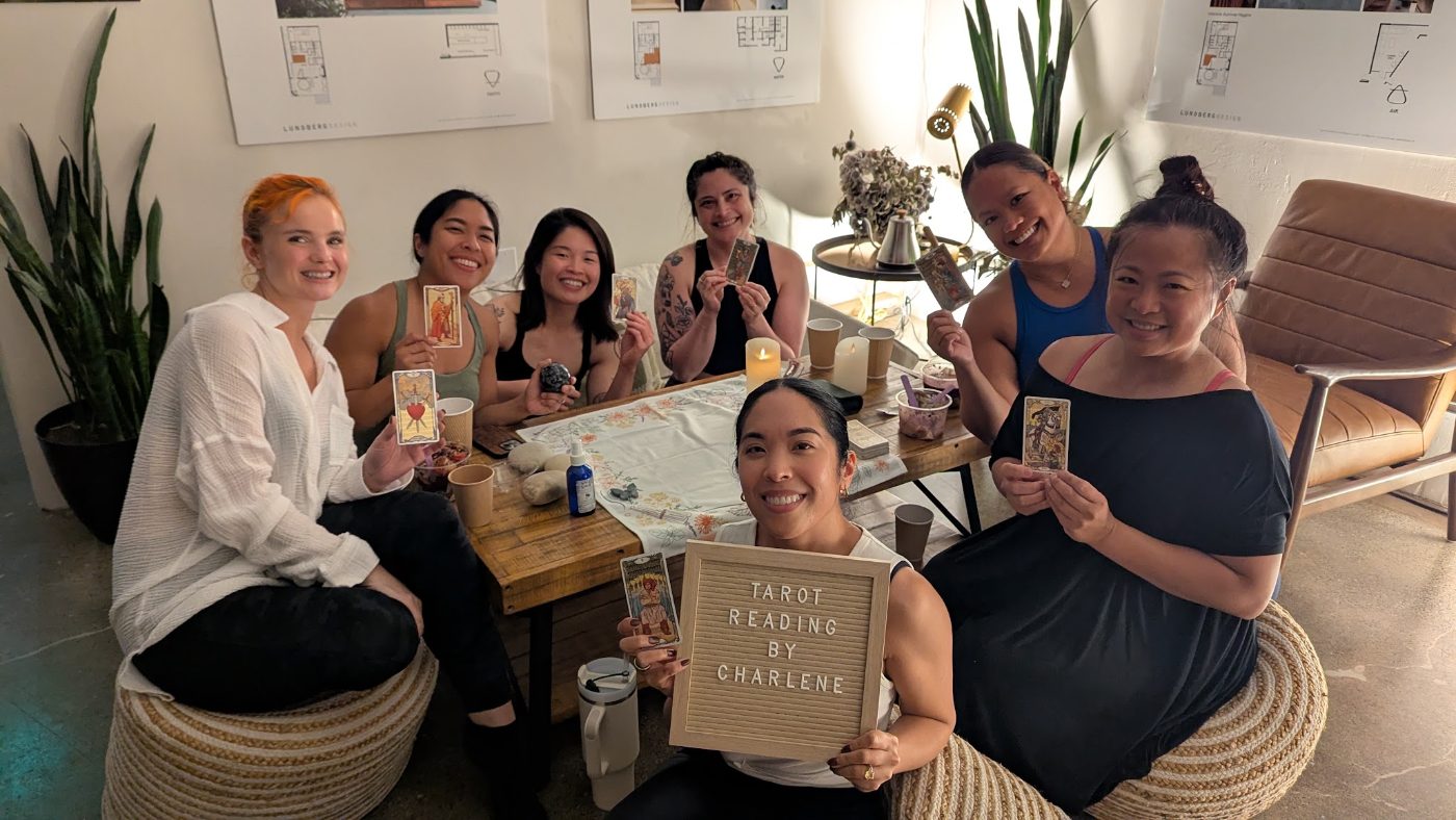 Seven smiling women around a tarot table at The Reset Retreat, each holding up a card, with Charlene holding a letterboard reading Tarot Reading by Charlene