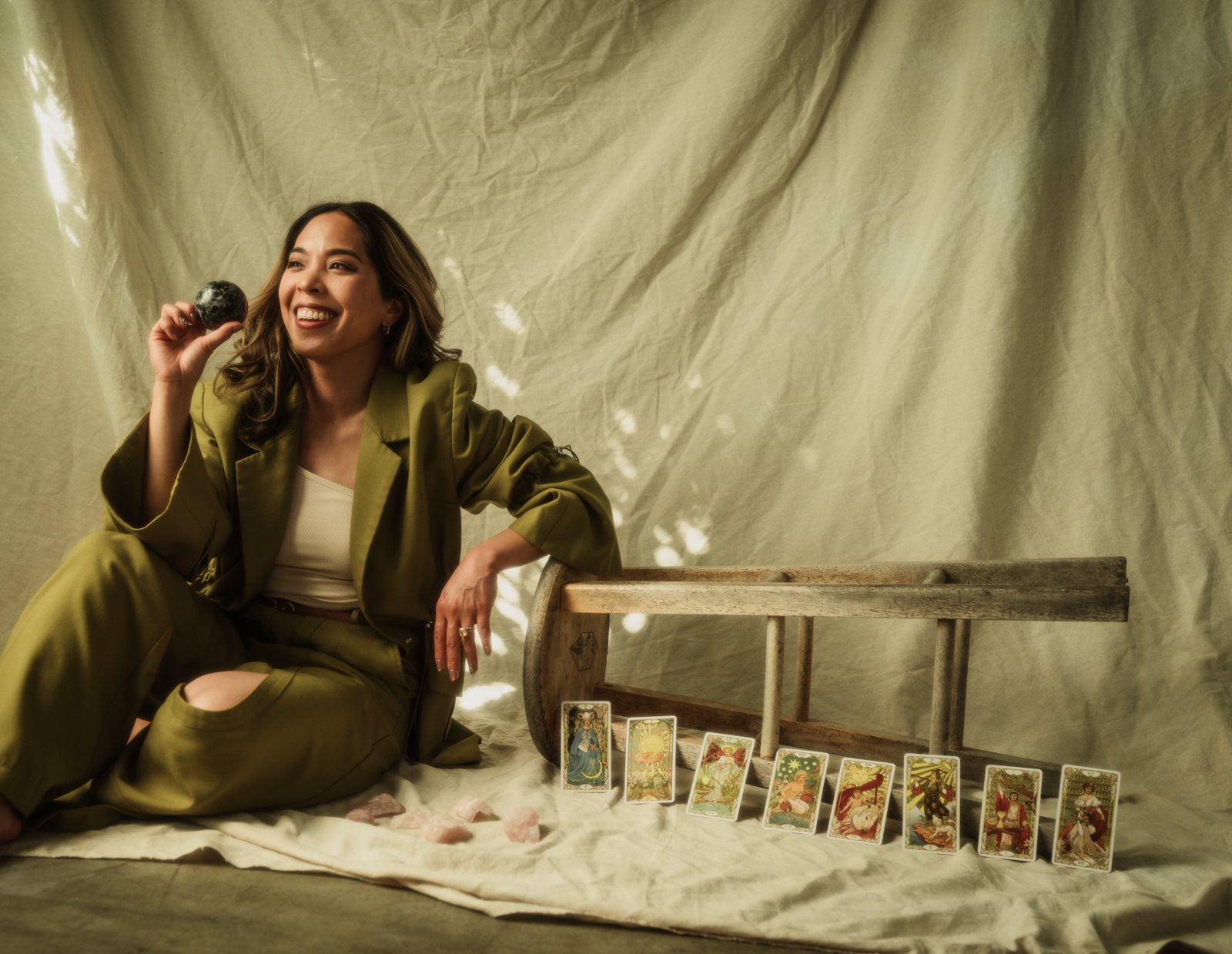 Charlene Platon smiling in an olive suit, seated in dappled daylight with tarot cards and rose quartz arranged beside her